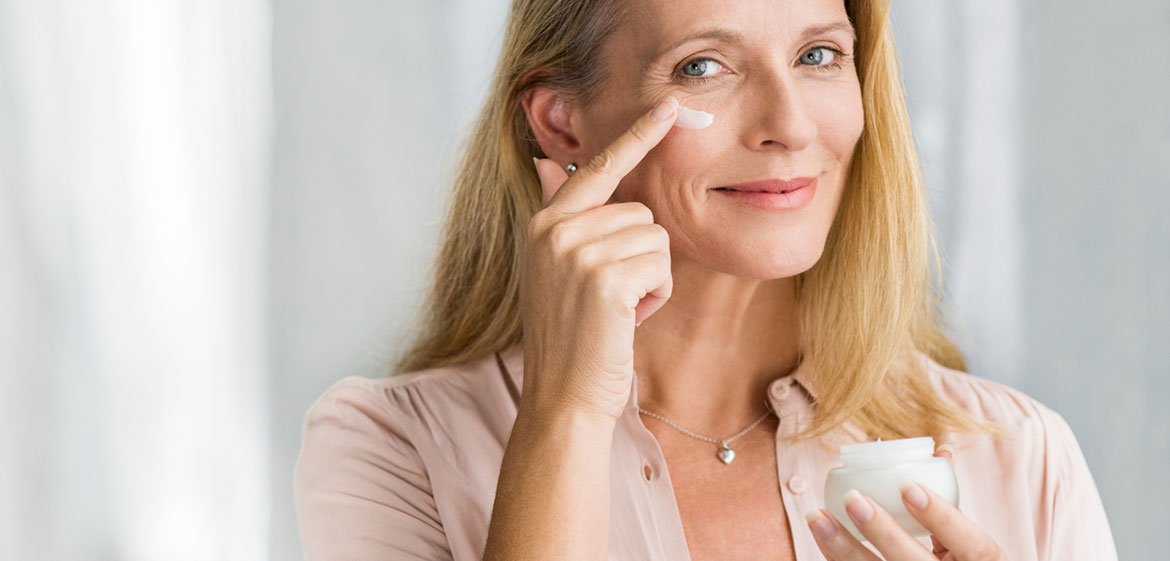 Eine ältere Frau mit langen blonden Haaren und grauem Haaransatz hält in der linken Hand einen Cremetiegel. Mit dem rechten Zeigefinger trägt sie die Creme auf ihr rechtes Jochbein auf.